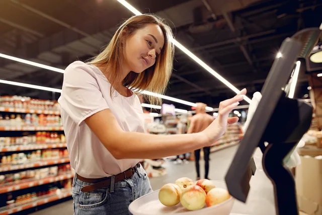 Side view of young woman in trendy clothes weighing peaches on scales while  shopping in supermarket during purchase food. Planning digital screen placements around shopper attention
